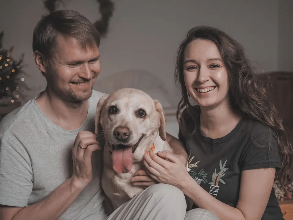 A couple sitting close on their bed with a grinning yellow Labrador between them.