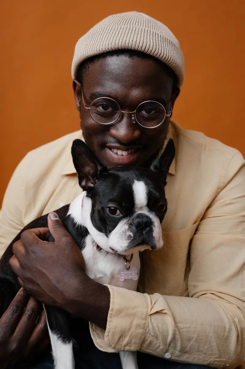 A man in a knit cap hugging a Boston terrier against an orange wall, both happy.